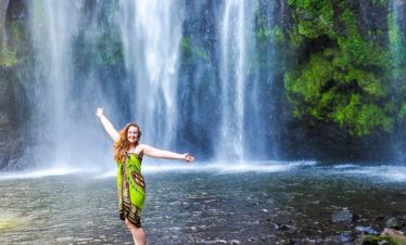 A visitor in a bright green dress stands with arms out by Materuni Waterfalls, where powerful water cascades through lush green foliage.