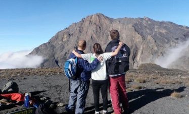 Three hikers standing together with arms around each other, overlooking the Meru Hills landscape.