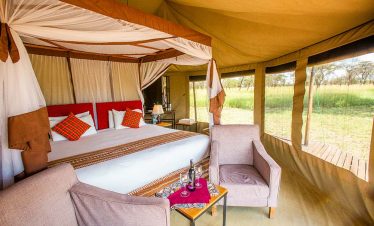 Inside Narudi tented camp, a canopy bed, lounge chairs and side table sit beside wide windows overlooking the safari landscape.