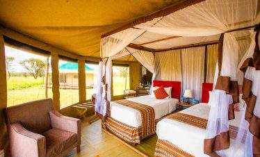 Inside Narudi Tented Camp, a safari tent suite shows twin beds with mosquito net canopies, warm earthy decor, and open views across the savanna.
